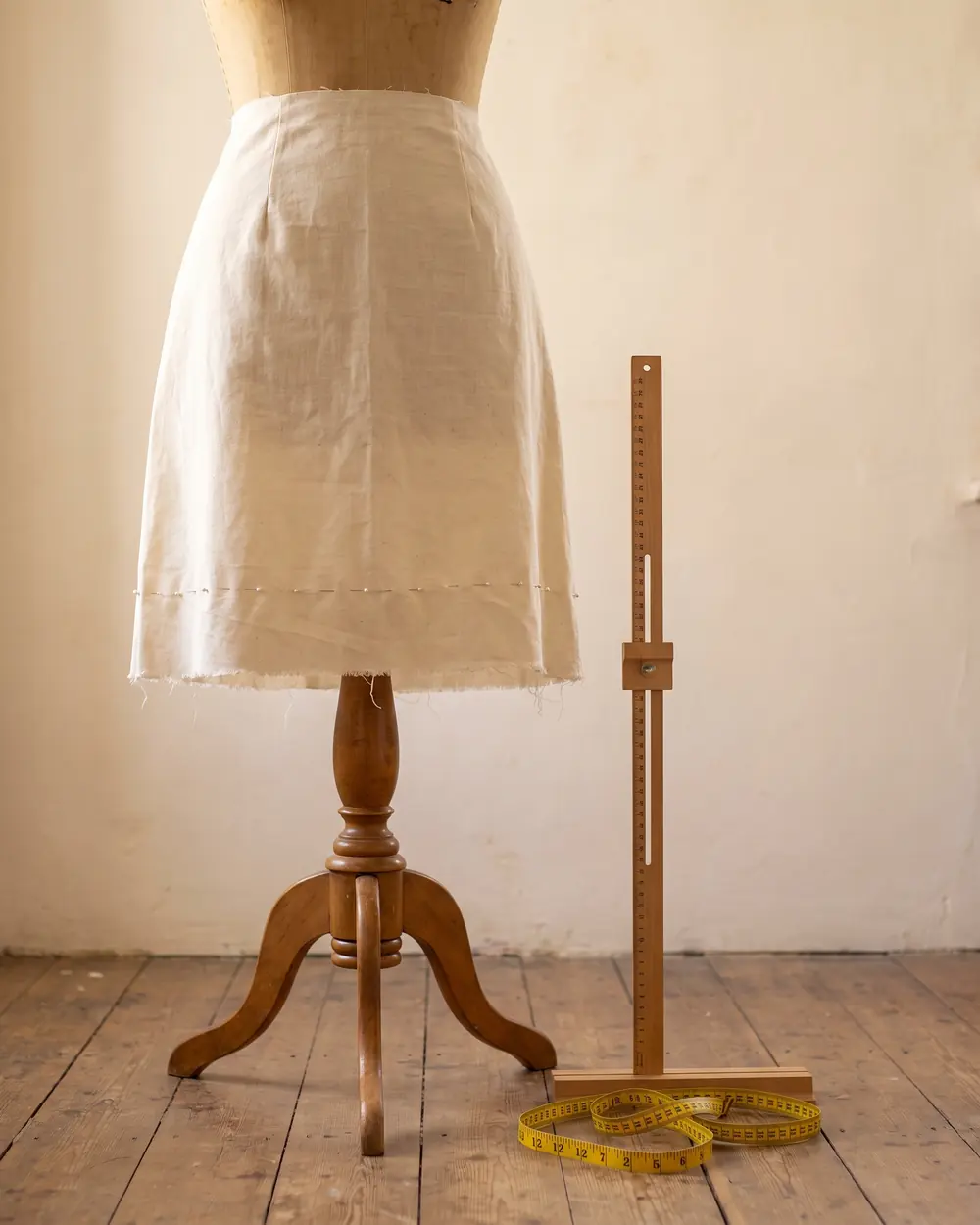 A muslin skirt toile on a dress form with a row of pins marking a new hem, beside a wooden hem marker