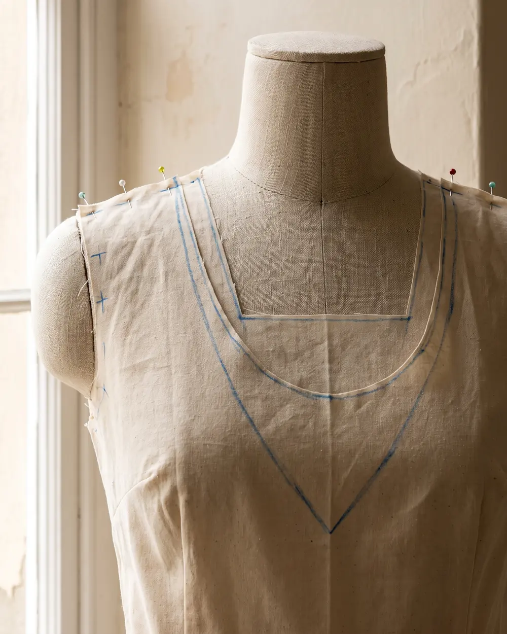 A muslin bodice on a dress form with a V, a scoop and a square neckline drawn in blue tailor's chalk